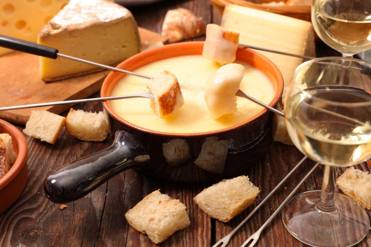 Traditionelles Schweizer Käsefondue in einem schwarzen Keramiktopf, in dem Brotwürfel an langen Gabeln in geschmolzenen Käse getaucht werden. Umgeben von Käsestücken, Brot, zwei Gläsern Weißwein und rustikalem Holzuntergrund.