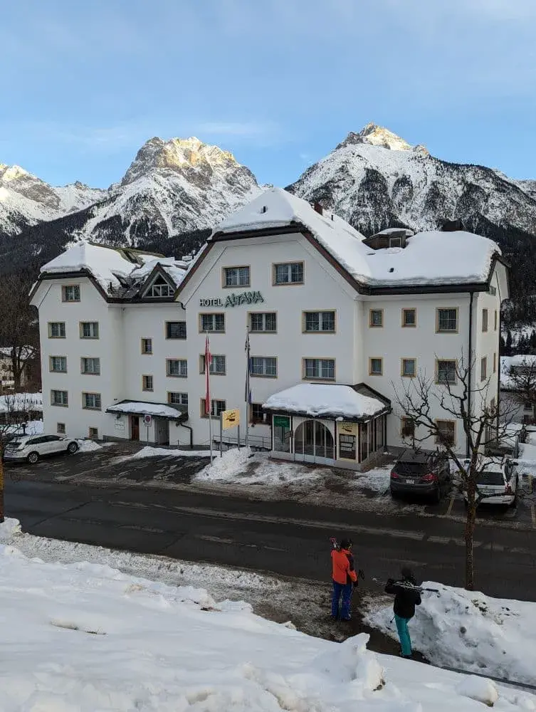 Hotel Atena in Scuol, mit Skifahrern davor und mit schneebedeckten Berggipfeln dahinter