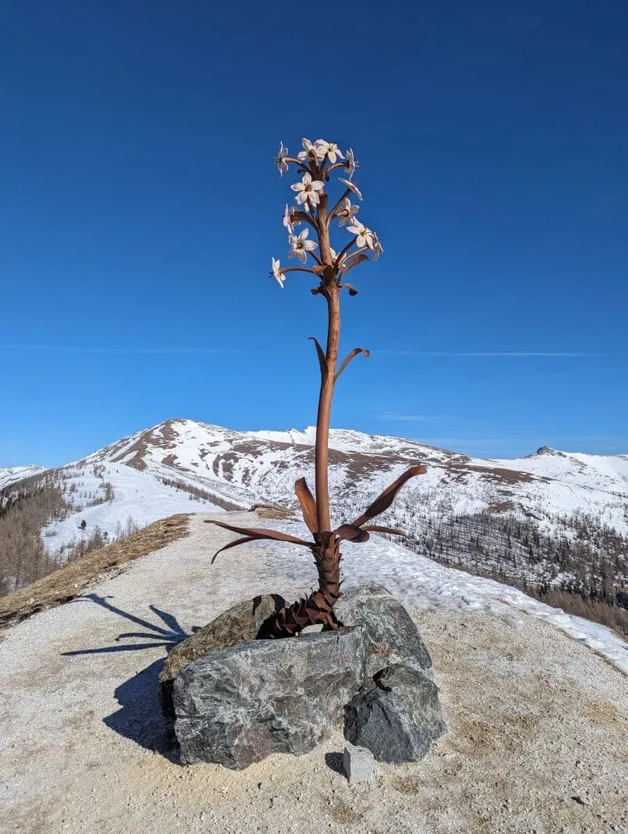 Eine Blume aus Eisen als Kunstwerk auf einem Aussichtspunkt nah der Biosphärenparkbahn Brunnach. Drumherum eine verschneite Winterlandschaft