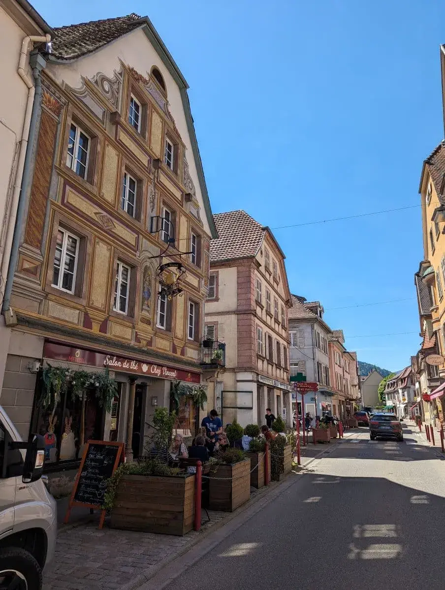 Die Grand Rue in Münster im Elsass, gesäumt von historischen Gebäuden. Menschen sitzen in einem Straßencafé und genießen das sonnige Wetter.