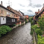 Ein malerischer Fluss fließt durch Kaysersberg, einem Dorf in der Umgebung von Colmar, Frankreich, gesäumt von traditionellen Fachwerkhäusern und bunten Gebäuden mit Holzbalkonen. Die Ufer des Flusses sind dicht mit grüner Vegetation bewachsen, und im Vordergrund sind blühende Blumen in einem kleinen Garten zu sehen.
