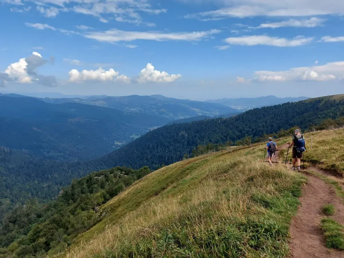Jan und mein Onkel mit Rucksäcken und Wanderstöcken gehen auf einem schmalen Pfad entlang eines Bergrückens hoch über dem Münstertal. Die Landschaft bietet weite Ausblicke auf bewaldete Hügel und Täler unter einem teilweise bewölkten blauen Himmel.
