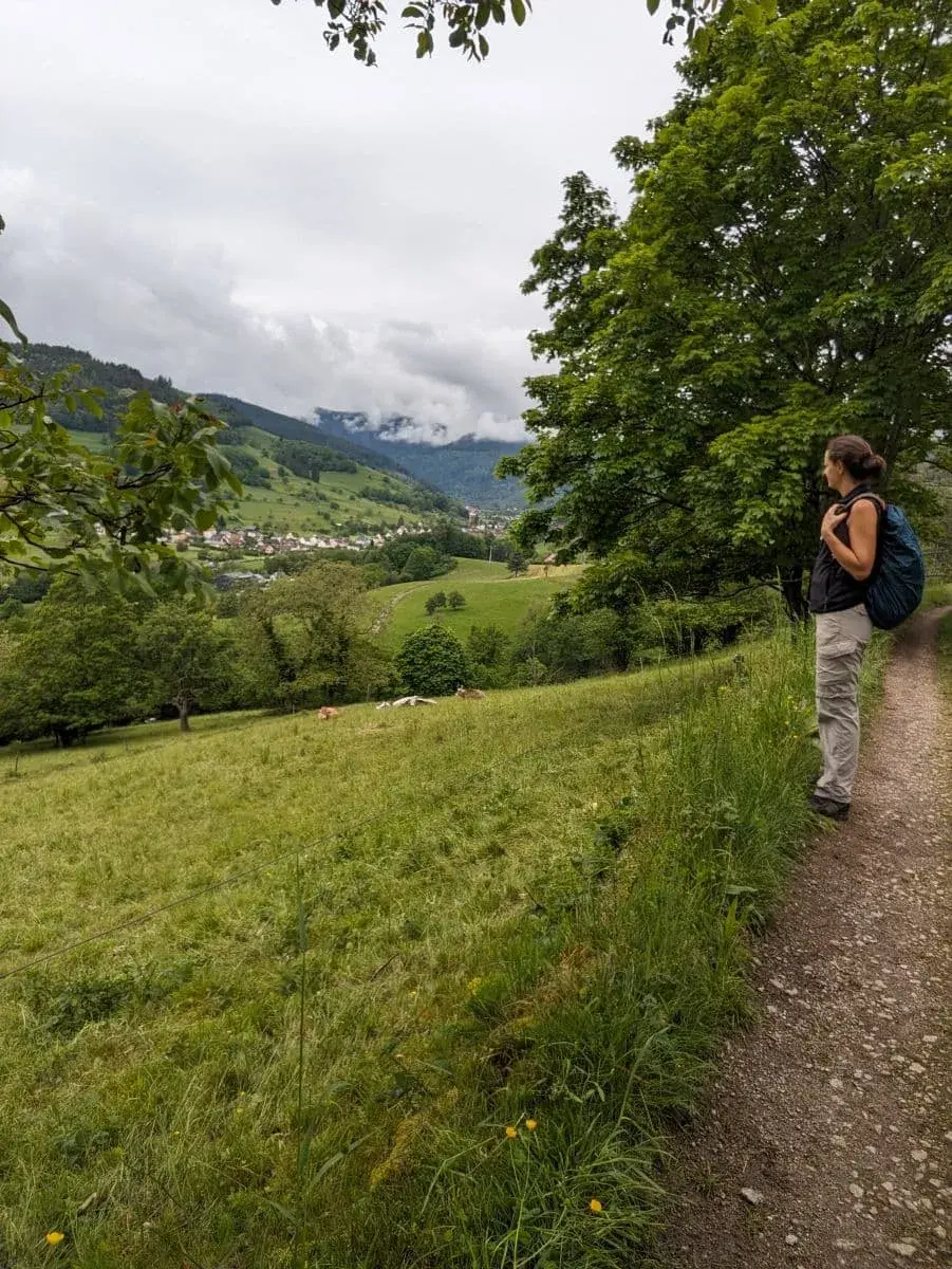 Linda mit einem Rucksack wandert auf einem Pfad im Münstertal. Die Landschaft ist grün und hügelig, mit Blick auf die Stadt Münster im Hintergrund und Wolken, die über die Berge ziehen.
