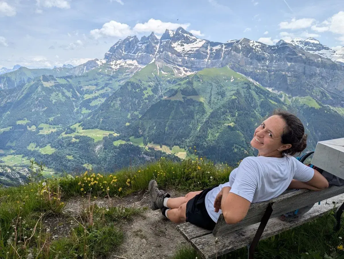 Linda sitzt auf einer Holzbank auf dem Gipfel des Croix du Culet und lächelt in die Kamera, umgeben von einem atemberaubenden Panorama aus grünen Tälern, blühenden Wiesen und schneebedeckten Gipfeln der Dents du Midi in den Alpen. Im Hintergrund erstrecken sich imposante Berge unter einem leicht bewölkten Himmel.