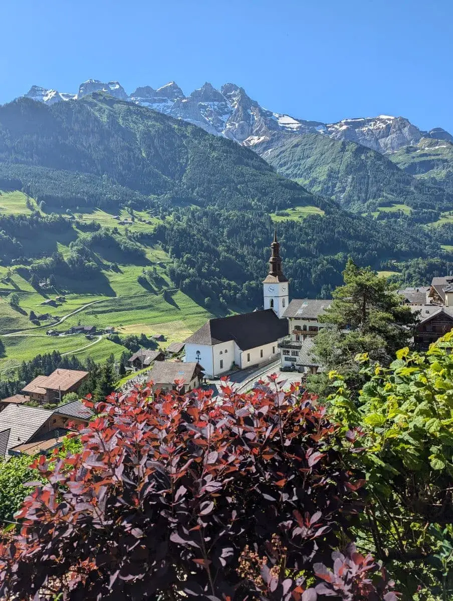 Eine malerische Ansicht der Dorfkirche von Val d'Illiez im Vordergrund, eingerahmt von bunten Sträuchern. Im Hintergrund erhebt sich majestätisch das Dents du Midi Gebirge, das mit schneebedeckten Gipfeln und grünen Hängen die Szene dominiert.