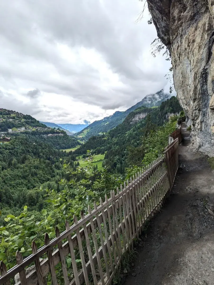 Ein schmaler Wanderweg mit einem Holzzaun verläuft entlang einer steilen Felswand und bietet einen weiten Blick auf das grüne Tal und die umliegenden Berge unter einem bewölkten Himmel.