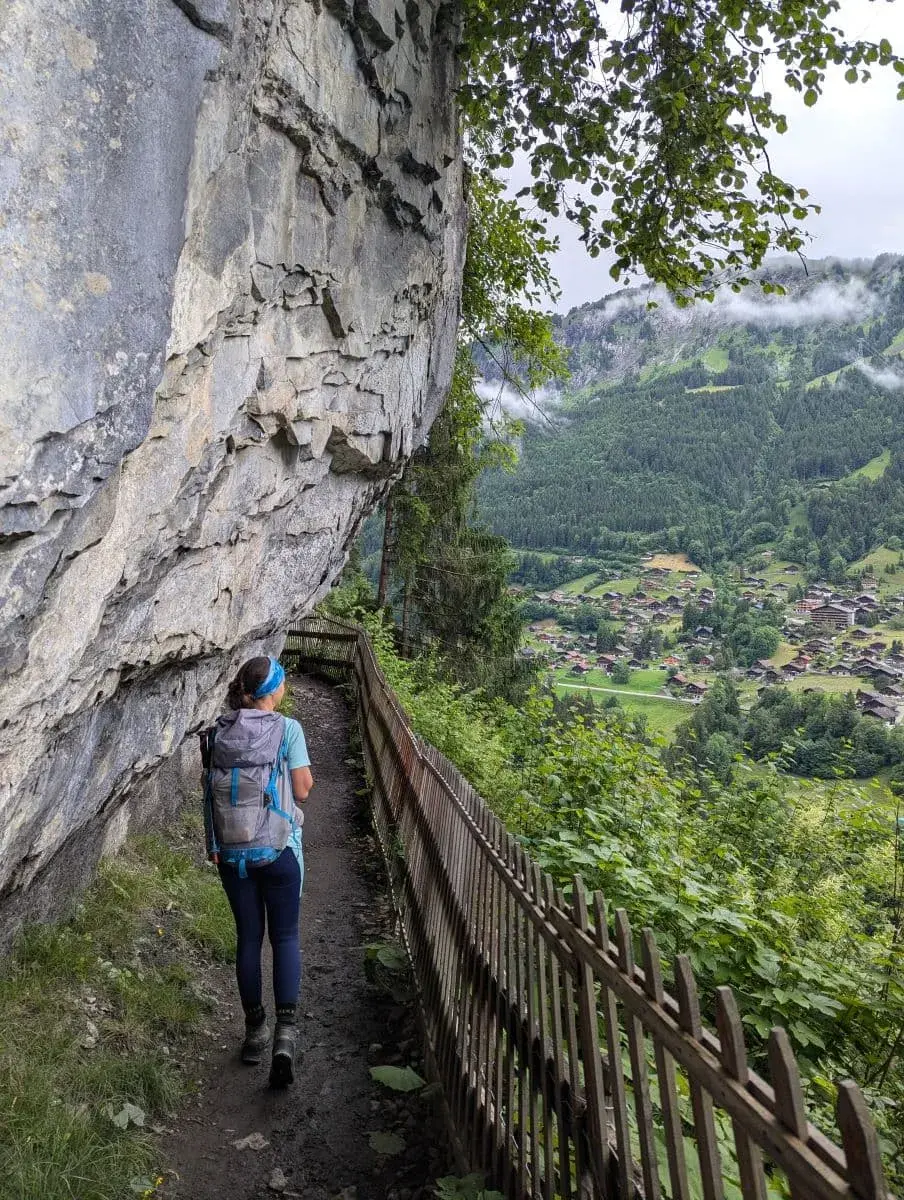Linda mit einem Rucksack wandert entlang eines schmalen Pfades, der von einem Holzzaun und einer steilen Felswand gesäumt ist, mit einem Panoramablick auf ein grünes Tal und Dörfer in der Ferne.