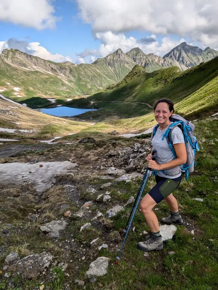 Linda mit Wanderstöcken und Rucksack steht vor einer malerischen Landschaft mit grünen Bergen und dem klaren Bergsee Lac de Chésery. Der Himmel ist leicht bewölkt, und die Umgebung zeigt einige Schneereste auf den Berghängen.