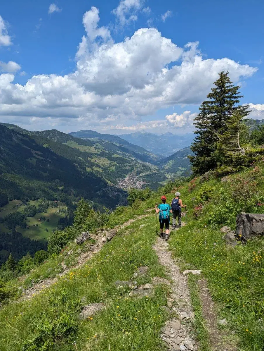 2 Wanderer laufen auf einem aussichtsreichen Hangweg auf das Bergdorf Champery zu. Man hat einen weiten Blick ins Tal, unter einem sonnigen, blauen Himmel.