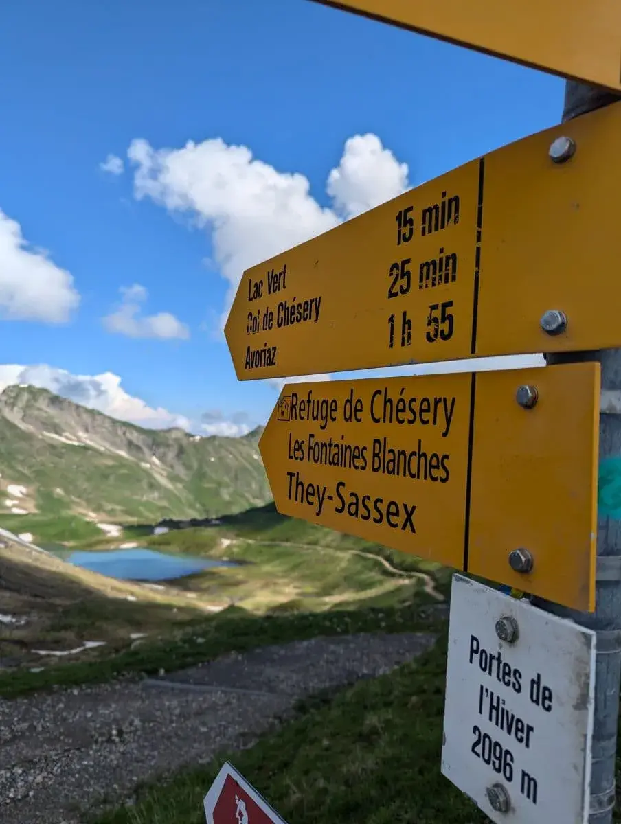 Gelbe Wegweiser auf einer Bergwiese beim Wandern in Champery zeigen die Richtungen zu verschiedenen Wanderzielen an, darunter Lac Vert, Col de Chésery und Avoriaz. Im Hintergrund sind grüne Berge und ein klarer See unter einem blauen Himmel zu sehen.