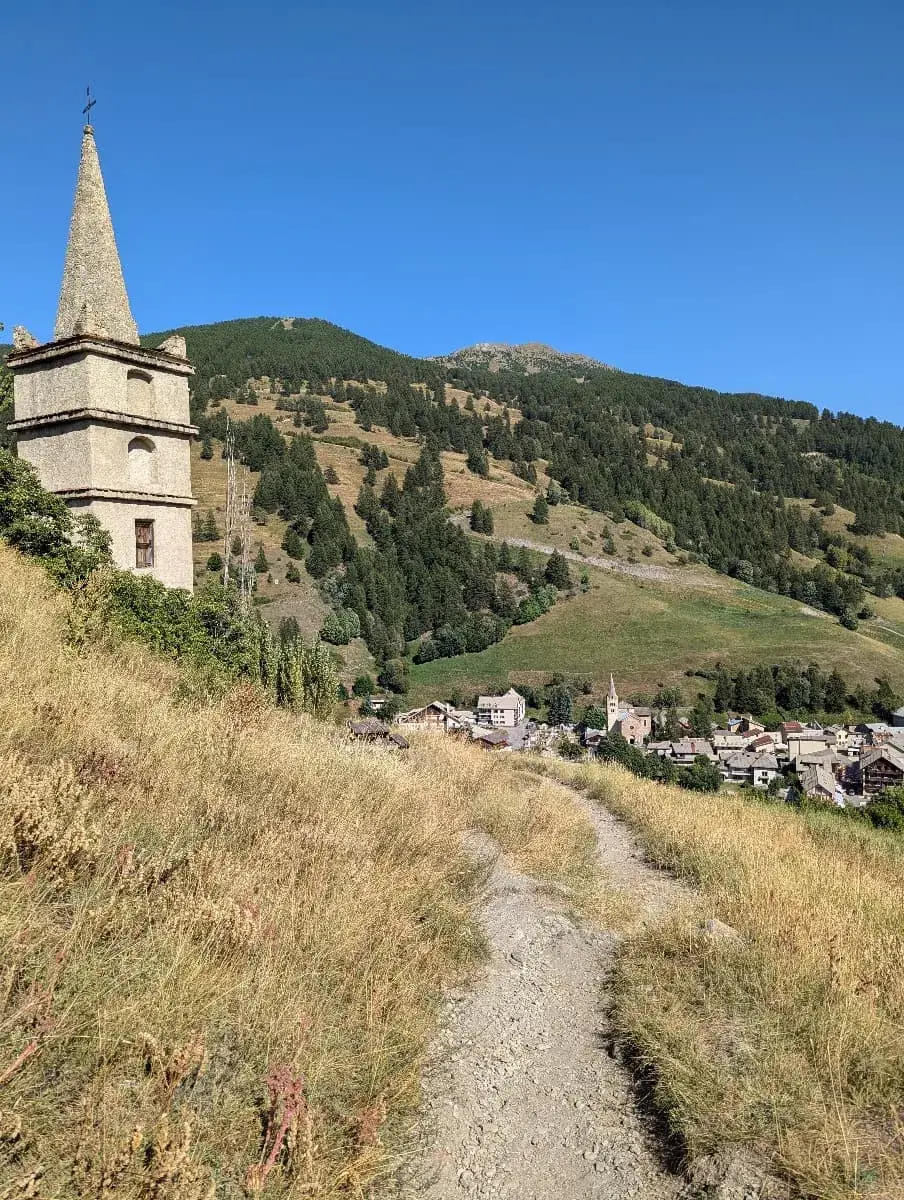 Ein schmaler Wanderweg führt durch eine sonnenverwöhnte Wiesenlandschaft, vorbei an einem alten Kirchturm mit spitzem Dach. Im Hintergrund liegt das Dorf Abries, eingebettet in grüne Hügel und bewaldete Berghänge unter einem strahlend blauen Himmel.
