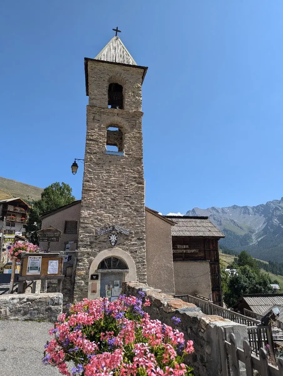Ein historischer Kirchturm aus Stein in Saint Veran, mit einem einfachen Kreuz an der Spitze und zwei Bögen, in denen Glocken hängen. Vor der Kirche ist ein Blumenbeet mit farbenfrohen Blumen zu sehen, die einen schönen Kontrast zu den rauen Steingebäuden und dem klaren, blauen Himmel bilden. Im Hintergrund erheben sich die Alpen des Queras majestätisch.