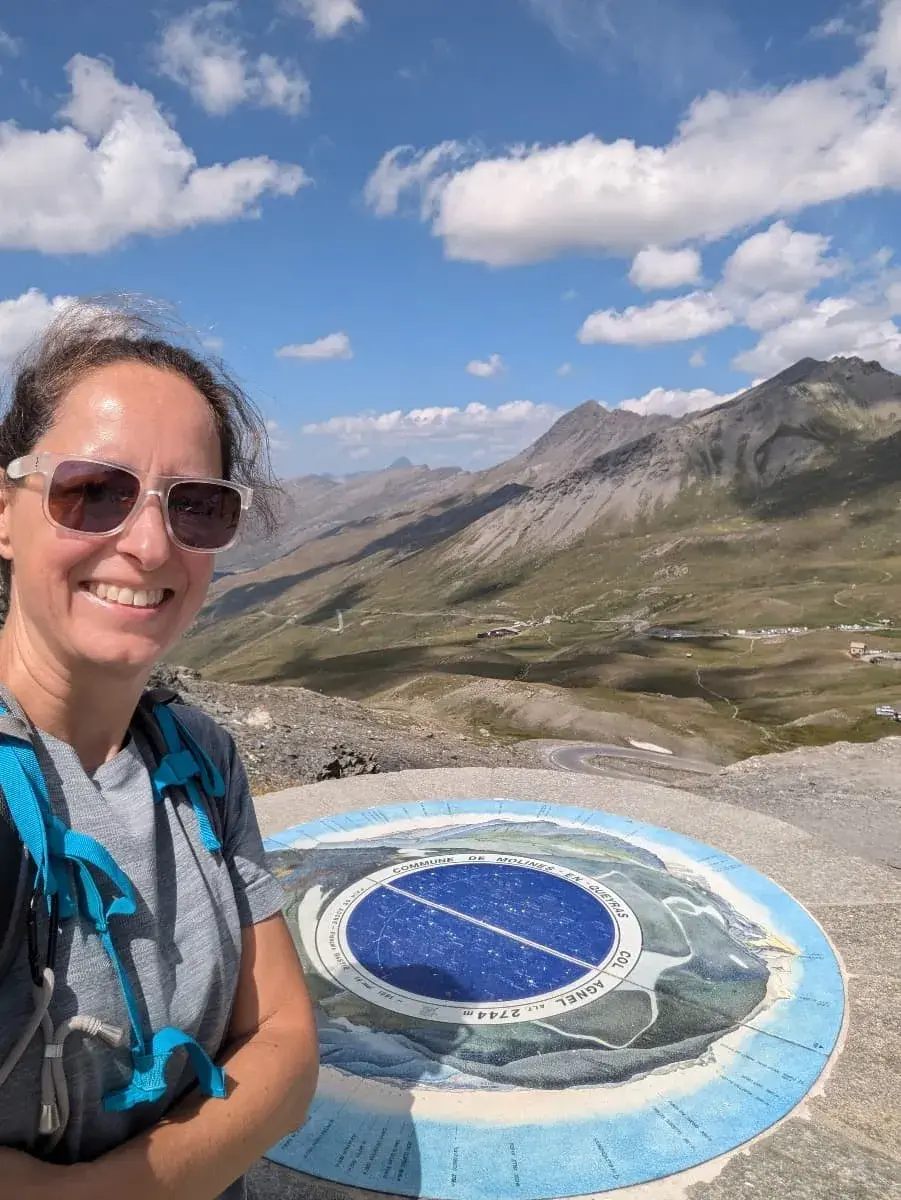 Linda steht lächelnd für ein Selfie auf dem Col Agnel, mit einem Panoramablick über die Alpen im Hintergrund. Vor ihr ist eine große runde Tafel zu sehen, die geografische Informationen über den Pass zeigt. Die umliegenden Berge und Täler erstrecken sich unter einem teils bewölkten, blauen Himmel, während die kurvigen Straßen in der Ferne sichtbar sind.