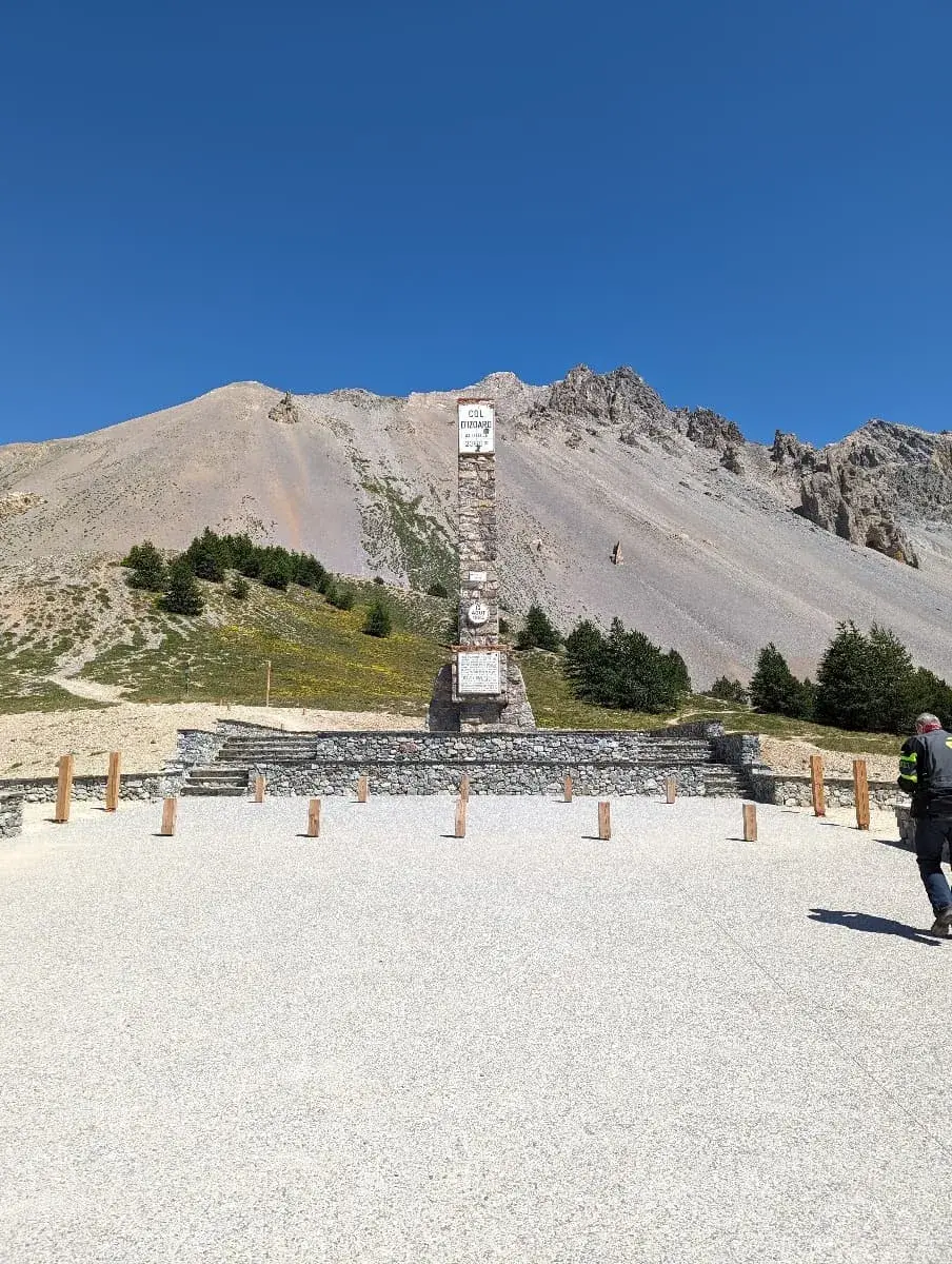 Ein steinernes Denkmal steht auf dem Gipfel des Col d'Izoard, einem bekannten Gebirgspass. Das Monument ist hoch und trägt Beschriftungen sowie Schilder, die den Namen und die Höhe des Passes anzeigen. Im Hintergrund erhebt sich eine karge, felsige Berglandschaft unter einem wolkenlosen, tiefblauen Himmel. Auf der rechten Seite des Bildes ist ein Mann in Motorradkleidung zu sehen, der vom Denkmal weggeht.