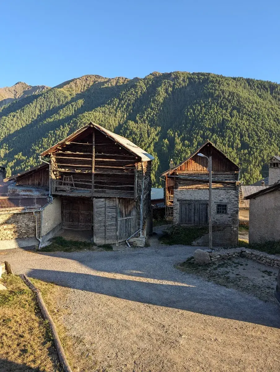 Zwei alte, traditionelle Holzhäuser stehen im sanften Licht des Spätnachmittags, umgeben von einer malerischen Berglandschaft. Die Schatten der nahen Berge zeichnen sich auf den grünen Hängen ab, während die Sonne den Himmel in warmes Licht taucht.