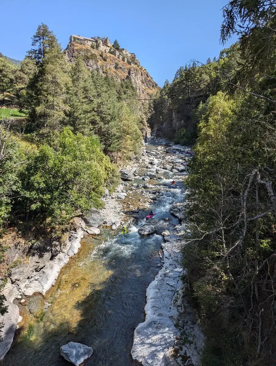 Ein kleiner, felsiger Fluss, der sich durch ein bewaldetes Tal schlängelt, mit einigen Kajakfahrern, die das wilde Wasser befahren. Hoch oben auf einem Felsen thront das beeindruckende Fort Queyras, das über das Tal blickt. Der klare Himmel und das dichte Grün der Bäume verleihen der Szene eine natürliche, alpine Schönheit.