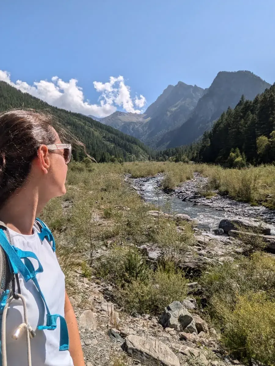 Linda blickt auf den Fluss Guil, der sich durch eine malerische Landschaft schlängelt, umgeben von dichten Wäldern und hohen, zerklüfteten Bergen. Der klare blaue Himmel mit vereinzelten Wolken vervollständigt die beeindruckende Alpenkulisse, die eine friedliche und natürliche Atmosphäre vermittelt.