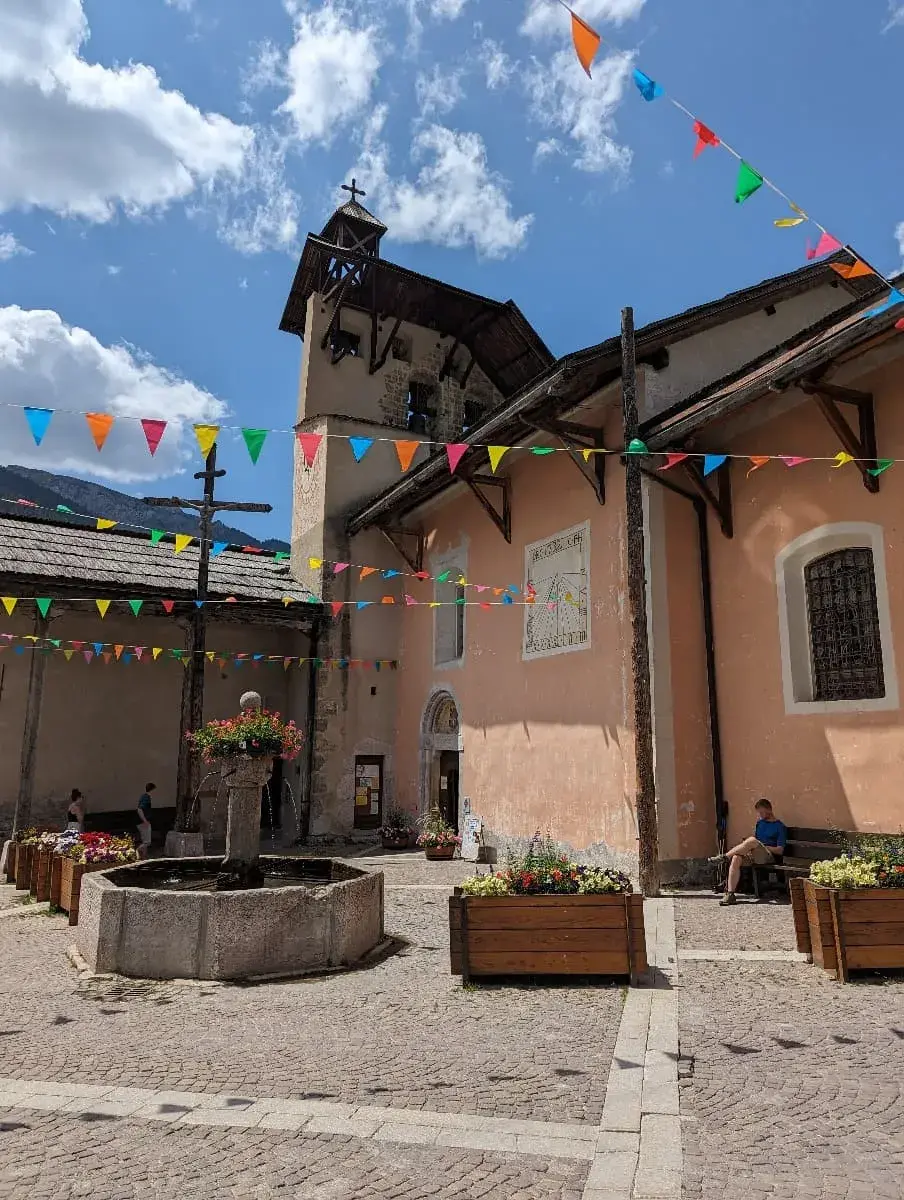 Ein sonniger Dorfplatz mit einer Kirche und bunten Wimpeln, die im Wind wehen. Blumenbeete und ein großer Brunnen in der Mitte des Platzes bringen Farbe in die Szene, während sich ein paar Menschen im Schatten entspannen.