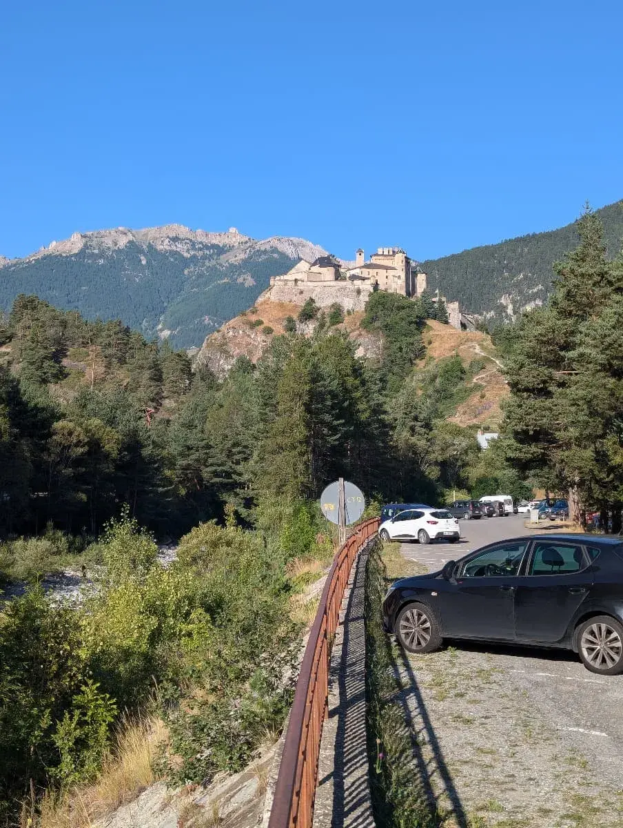 Das Fort Queyras, ein imposantes historisches Gebäude, sitzt majestätisch auf einem Hügel und überblickt eine bewaldete Landschaft. Im Vordergrund sind ein Parkplatz und ein Geländer zu sehen, während die Festung vor einem klaren, strahlend blauen Himmel steht. Die steinernen Mauern und Türme des Forts wirken fest und schützend.