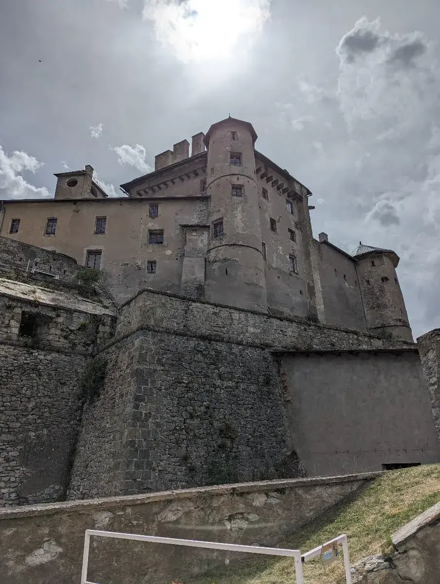 Eine massive, historische Festung, das Fort Queyras, erhebt sich imposant auf einem Felsvorsprung. Die dicken Steinmauern und hohen Türme stehen gegen einen bewölkten Himmel, durch den die Sonne dramatisch hindurchscheint. Die Festung wirkt robust und wehrhaft, typisch für militärische Bauwerke vergangener Zeiten.