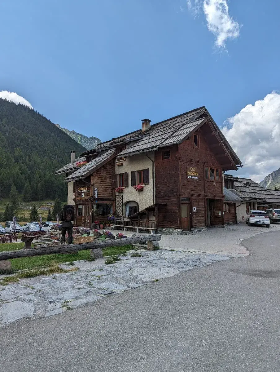 Ein rustikales Gasthaus aus Holz, bekannt als "Gîte des Rabious", liegt in einem kleinen Bergdorf. Die blumengeschmückte Fassade und die umliegenden grünen Hügel fügen sich perfekt in die alpine Landschaft ein.