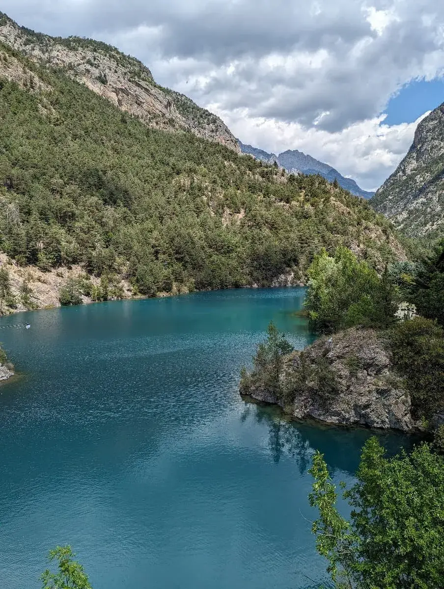 Ein malerischer Stausee, umgeben von dicht bewaldeten Hängen und steilen Berghängen. Das türkisfarbene Wasser reflektiert den teils bewölkten Himmel und bildet einen ruhigen Kontrast zu den felsigen Ufern und grünen Bäumen. Im Hintergrund sind die dramatischen Gipfel der Alpen des Queyras zu sehen.