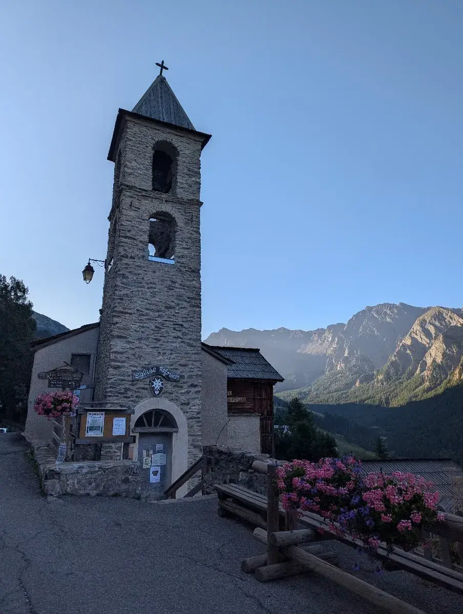 Eine alte Steinkirche mit einem Glockenturm steht im Vordergrund, während die Berge im Hintergrund von der Morgensonne beleuchtet werden. Der klare Himmel und die blühenden Blumen am Rand des Bildes sorgen für eine friedliche Atmosphäre.