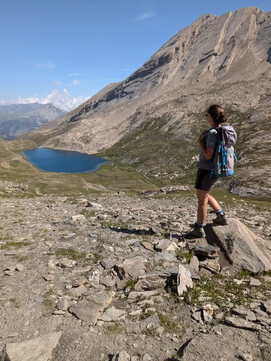 Linda steht auf einem Felsen und blickt auf den türkisblauen Lac Foreant, der in einem weiten Bergkessel auf dem GR58 liegt. Die steilen, felsigen Berghänge und der klare Himmel schaffen eine spektakuläre alpine Szenerie.