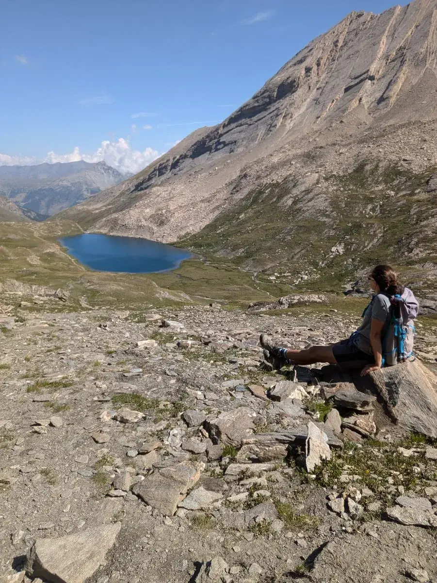 Linda sitzt auf einem großen Felsen und blickt auf den malerischen Lac Foréant, eingebettet in einer felsigen Alpenlandschaft. Der klare, blaue See liegt ruhig inmitten grüner Wiesen und steiler Bergflanken, die bis in den Himmel ragen. Der wolkenlose Himmel verstärkt das Gefühl von Ruhe und Abgeschiedenheit.