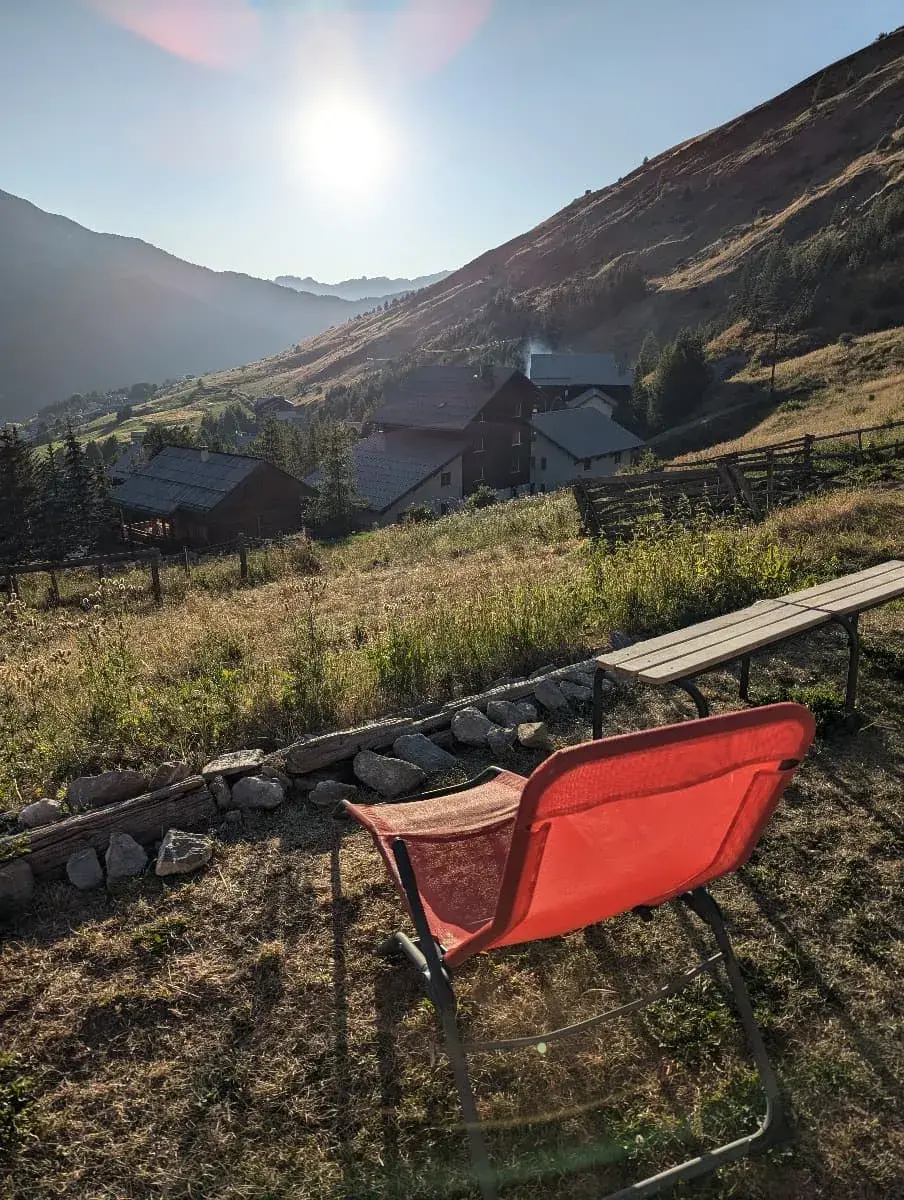 Ein roter Liegestuhl steht vor der Berglandschaft des Queyras, die im Licht der tiefstehenden Sonne badet. Die Szene wirkt friedlich und einladend, mit Blick auf das Tal und die umliegenden Berghänge, die im Dämmerlicht warm beleuchtet sind.