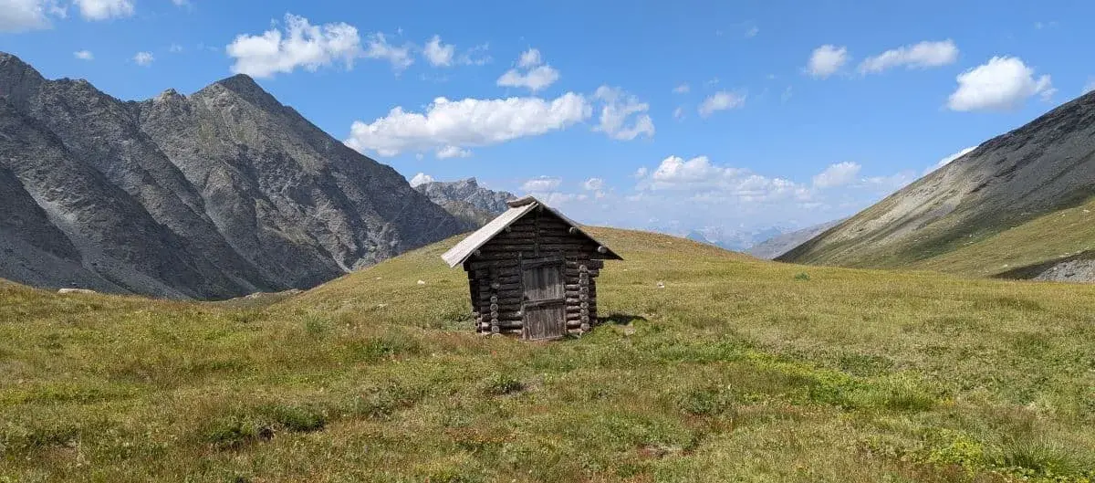 Eine abgelegene Holzhütte steht in einer weiten Bergwiese, umgeben von felsigen Bergen und strahlend blauem Himmel. Die Einsamkeit der Hütte und die umgebende Natur schaffen eine Atmosphäre von Ruhe und Abgeschiedenheit.