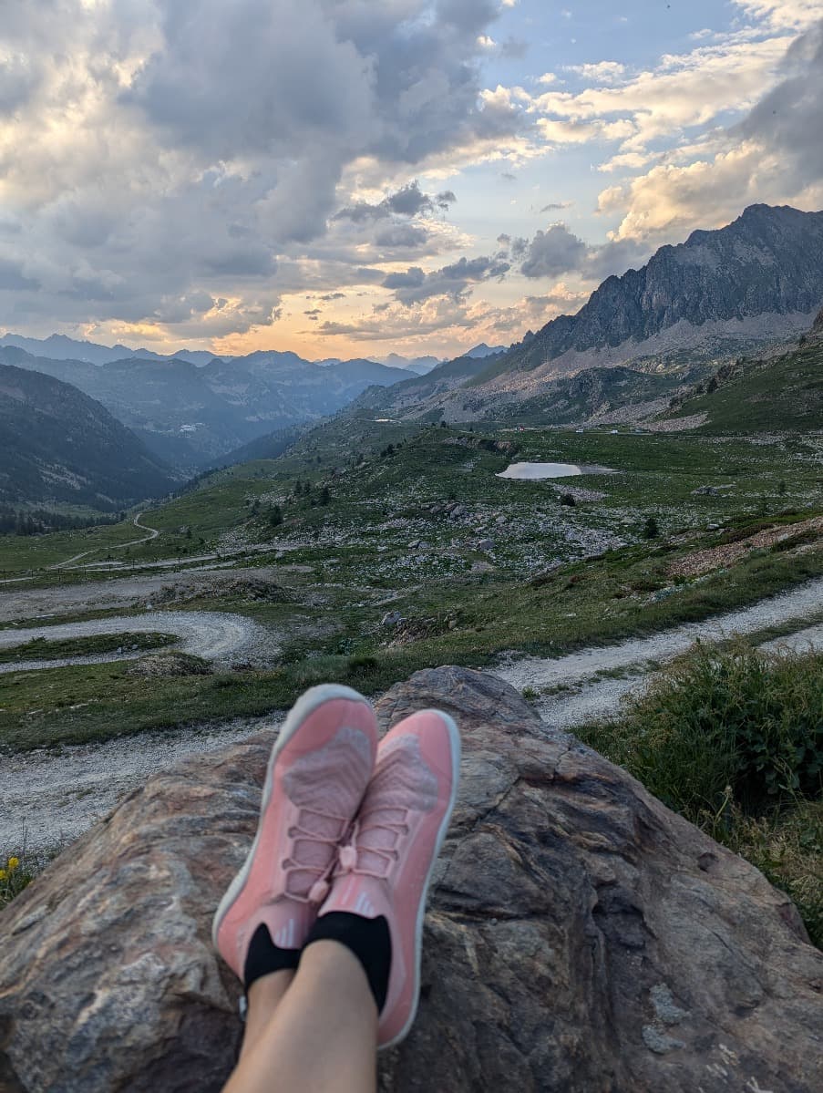 Nahaufnahme von Lindas pinkfarbenen Primus Trail Knit FG Schuhen, während sie auf einem Felsen sitzt und eine weitläufige Berglandschaft mit einem Serpentinenpfad und Wolken im Hintergrund betrachtet.