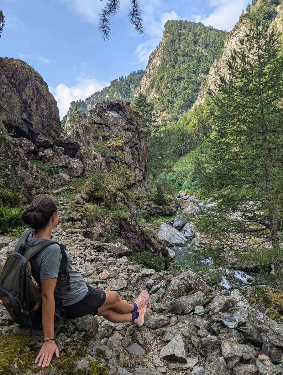 Linda sitzt auf einem steinigen Pfad nahe eines Baches, umgeben von hohen Felsen und grünen Bäumen. Sie trägt die Primus Trail Knit Schuhe, während sie die ruhige Umgebung genießt.