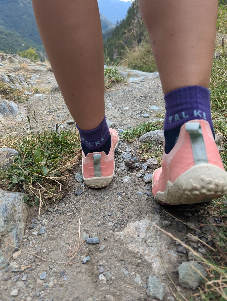 Nahaufnahme von Lindas Füßen, die auf einem steinigen Wanderpfad gehen. Sie trägt die pinkfarbenen Primus Trail Knit FG Schuhe und lila Socken von Falke