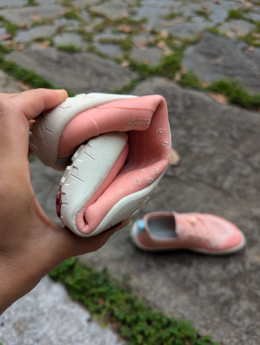 Linda hält einen eingerollten pinken Barfußschuh Primus Trail Knit FG in der Hand. Im Hintergrund liegt der zweite Schuh auf dem steinigen Boden