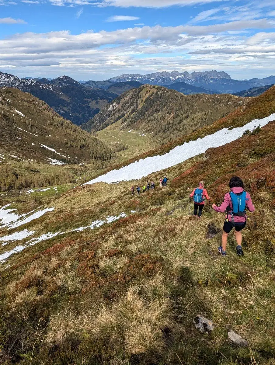 Eine Gruppe Wanderer bewegt sich einen grasbewachsenen Hang hinab, vorbei an schmelzendem Schnee. Die Landschaft ist von sanften Bergen und vereinzelten Schneefeldern geprägt, während der Weg durch eine malerische, leicht bewaldete Region führt. Im Hintergrund erheben sich die Berggipfel unter einem blauen Himmel mit leichter Bewölkung.