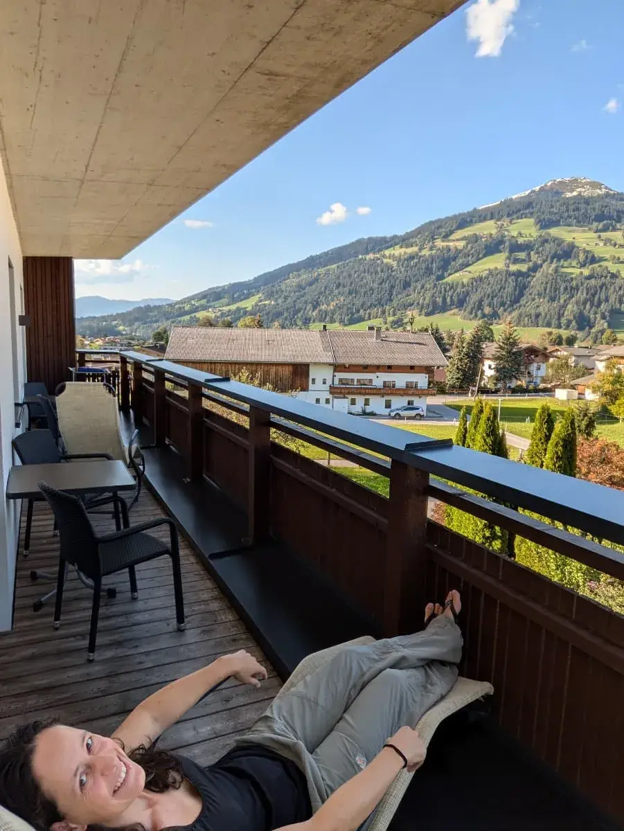 Linda liegt entspannt auf einem Balkonstuhl mit Blick auf die grüne Berglandschaft der Hohen Salve. Im Hintergrund sind traditionelle Tiroler Häuser und bewaldete Hügel zu sehen. Der Himmel ist klar mit einigen wenigen Wolken.