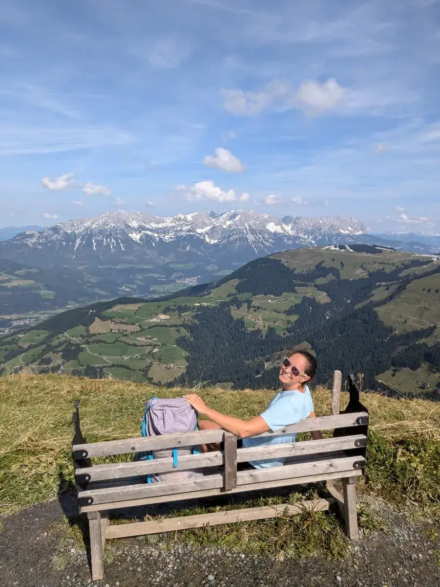 Linda sitzt entspannt auf einer Holzbank auf der Hohen Salve und blickt auf die schneebedeckten Gipfel des Wilden Kaisers im Hintergrund. Die Aussicht zeigt eine weitläufige Berglandschaft mit grünen Wiesen und Dörfern im Tal. Der klare Himmel unterstreicht die Weite der Tiroler Alpen. 