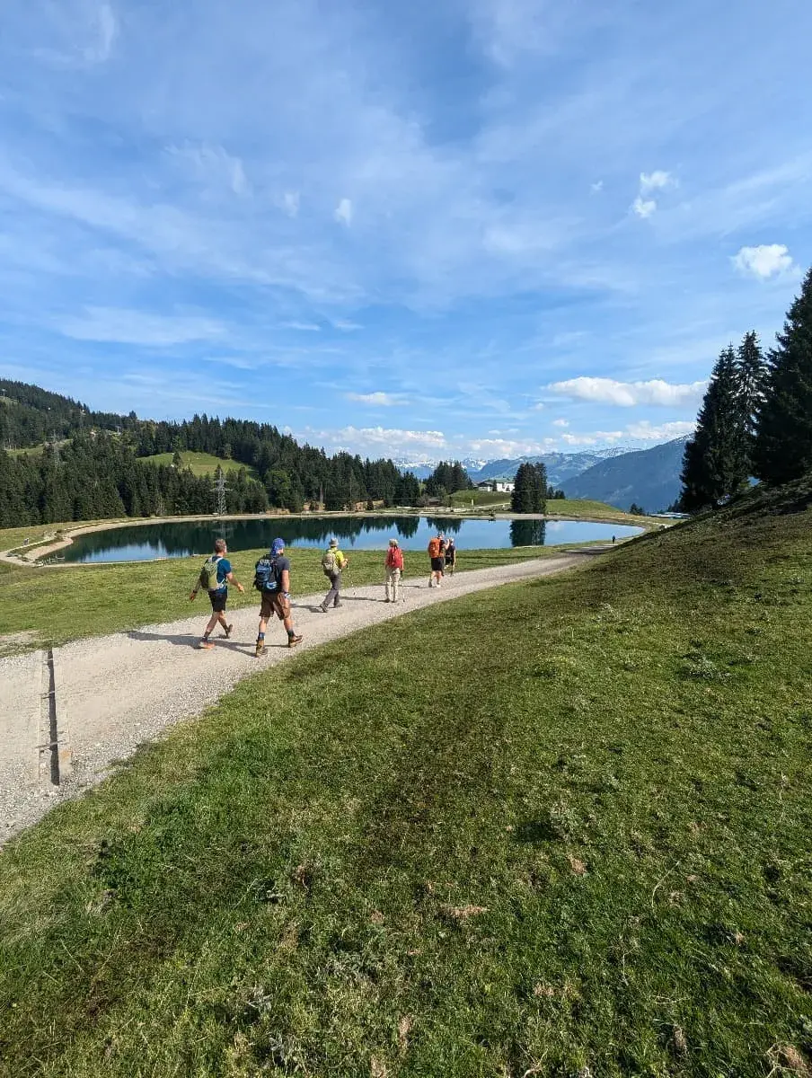 Eine Gruppe Wanderer geht entlang eines Schotterwegs, der am Filzalmsee vorbeiführt. Der Bergsee liegt ruhig inmitten einer hügeligen Landschaft mit saftig grünen Wiesen und Wäldern. Im Hintergrund ragen Berggipfel in den blauen Himmel. Ein idyllischer Ort zum Wandern im Brixental.