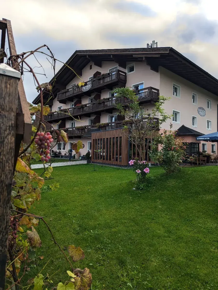 Ein traditionelles Alpenhaus mit mehreren Etagen und Holzbalkonen steht in einem gepflegten Garten. Im Vordergrund sind reife Trauben und blühende Pflanzen zu sehen, während das Gebäude im typischen alpenländischen Stil gehalten ist.