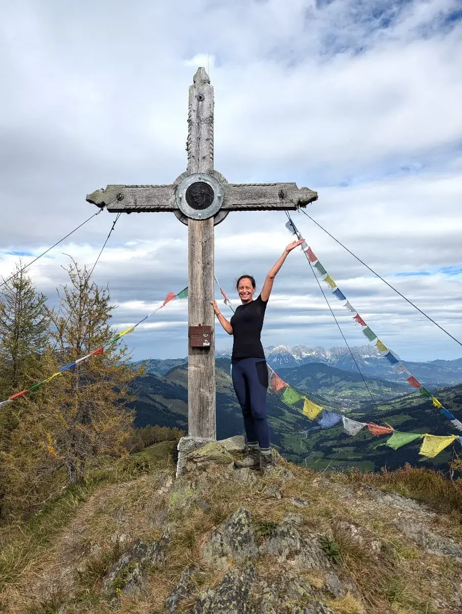 steht neben einem Gipfelkreuz, die Arme freudig in die Luft gestreckt. Um das Kreuz flattern bunte Gebetsfahnen, während im Hintergrund die beeindruckenden Alpen sichtbar sind. Die Wolkendecke ist locker und gibt den Blick auf die umliegenden Täler und Berge frei.
