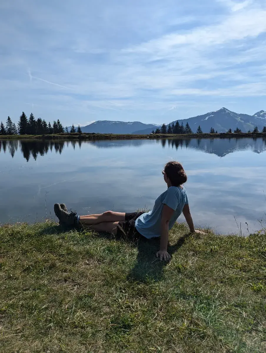 Linda sitzt am Ufer des klaren Hohe Salve Sees und genießt die Aussicht auf die umliegenden Kitzbüheler Alpen. Die ruhige Wasseroberfläche spiegelt den Himmel und die Berglandschaft wider. 