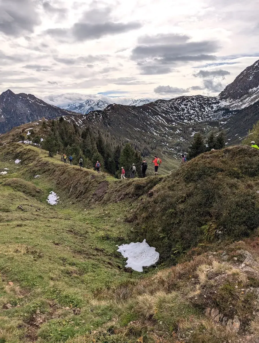 Wanderer gehen entlang eines schmalen Bergkamms, umgeben von leicht schneebedeckten Wiesen und Nadelbäumen. Der Pfad führt durch eine weitläufige Berglandschaft, während sich der Himmel mit Wolken bedeckt zeigt und das Licht sanft auf die Umgebung fällt.