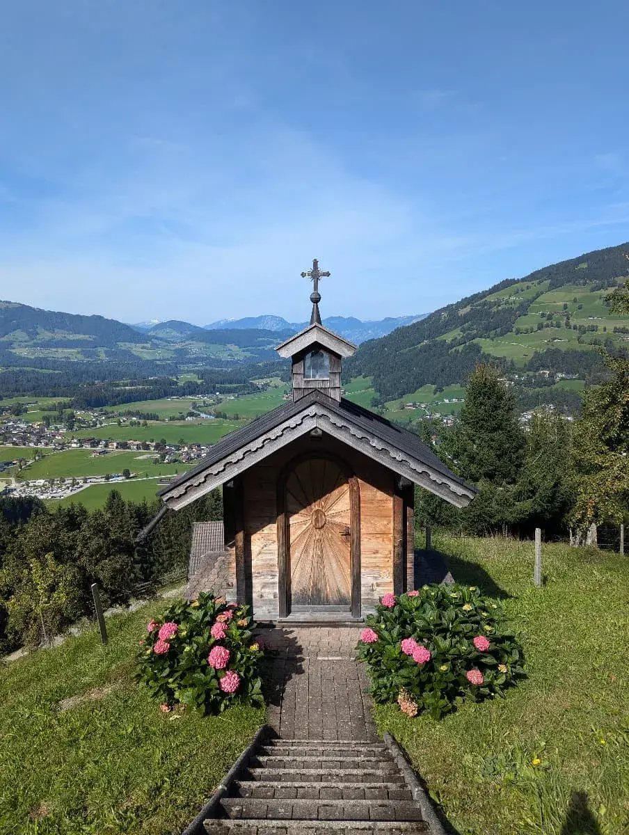 Eine kleine hölzerne Kapelle steht auf einer Anhöhe im Brixental, flankiert von blühenden Hortensienbüschen. Ein schmaler gepflasterter Weg führt hinauf, während sich im Hintergrund eine grüne Tal- und Berglandschaft erstreckt. 