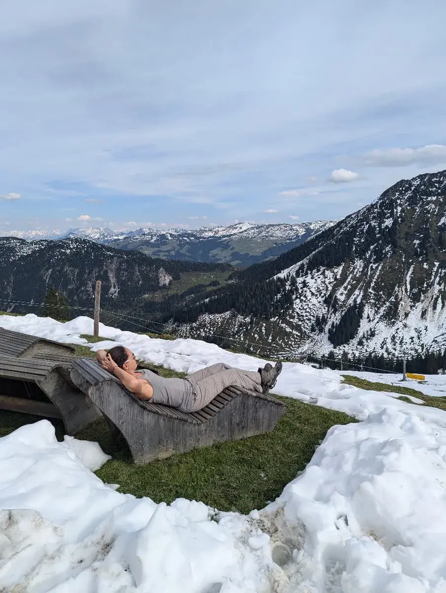 Linda liegt entspannt auf einem Holz-Liegestuhl, umgeben von schmelzendem Schnee auf einer Wiese. Im Hintergrund erstrecken sich die verschneiten Alpen des Brixentals unter einem klaren Himmel. Eine erholsame Pause beim Wandern im Brixental.