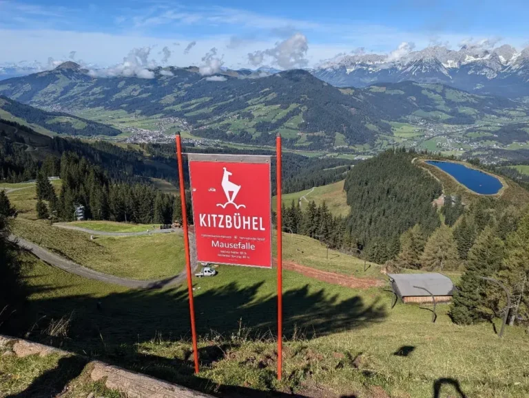Ein rotes Kitzbühel-Schild mit der Aufschrift „Mausefalle“ steht auf einer grünen Wiese mit einem weiten Ausblick auf die umliegenden Berge und Täler des Streif Wanderweges. Der Hintergrund zeigt eine beeindruckende Panoramasicht, einschließlich schneebedeckter Gipfel der Kitzbüheler Alpen und eines kleinen Sees.