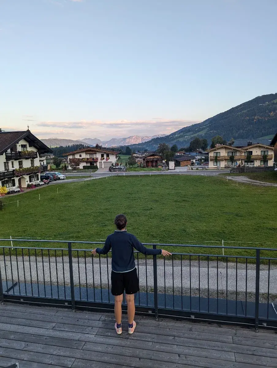 Linda steht auf einer Terrasse und blickt über eine große Wiese auf die umliegenden Berge und Dörfer. Die Sonne geht langsam unter, und die Berge im Hintergrund sind leicht vom Abendlicht erleuchtet. Die Atmosphäre wirkt ruhig und entspannend.