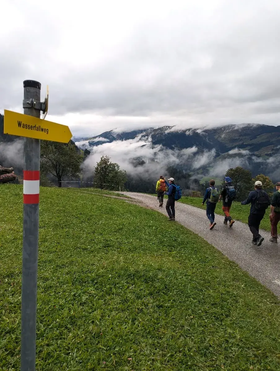 Eine Gruppe Wanderer folgt einem leicht ansteigenden Weg, der von einem Schild mit der Aufschrift „Wasserfallweg“ gekennzeichnet ist. Der Himmel ist bewölkt, und Nebelschwaden ziehen über die umliegenden Berge und Täler. Die grüne Wiese und der wolkenverhangene Hintergrund schaffen eine mystische Atmosphäre.