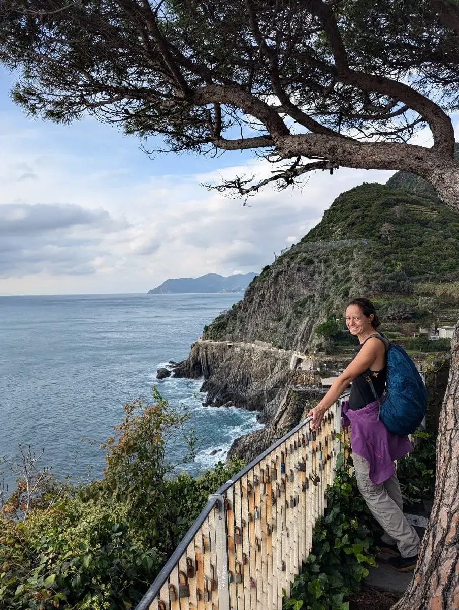 Linda steht auf einem mit Vorhängeschlössern behängten Geländer und genießt den Blick über das Meer und die felsige Küste bei Manarola, umrahmt von grünen Hügeln und einem Baum im Vordergrund.