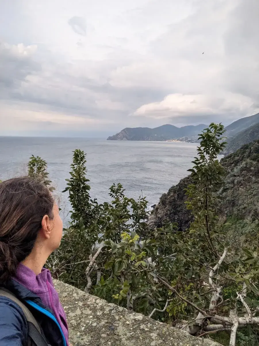 Linda schaut über eine Balustrade auf das Meer und die zerklüftete Küste von Corniglia im Cinque Terre Nationalpark. Ein paar bewaldete Felsen stehen auf den Klippen, das Meer ist rau und wellig.